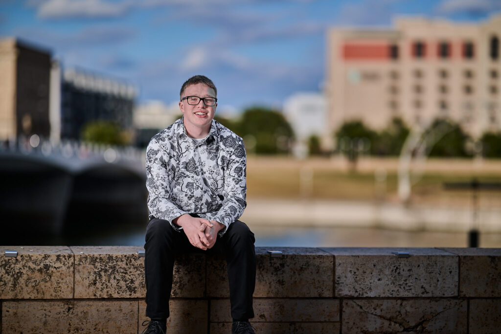 senior guy on a photoshoot, looking at the camera in downtown des moines, with the city in the background