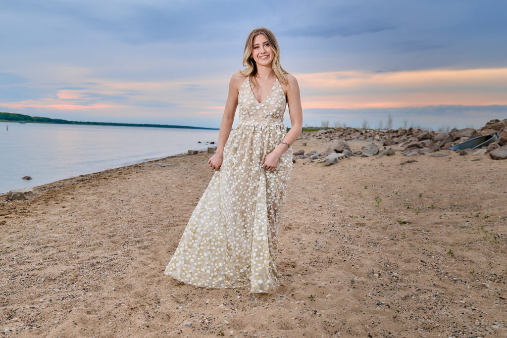 Senior girl session at Saylorville lake, with her in a beige dress, walking towards the camera, smiling with the sunset in the background.