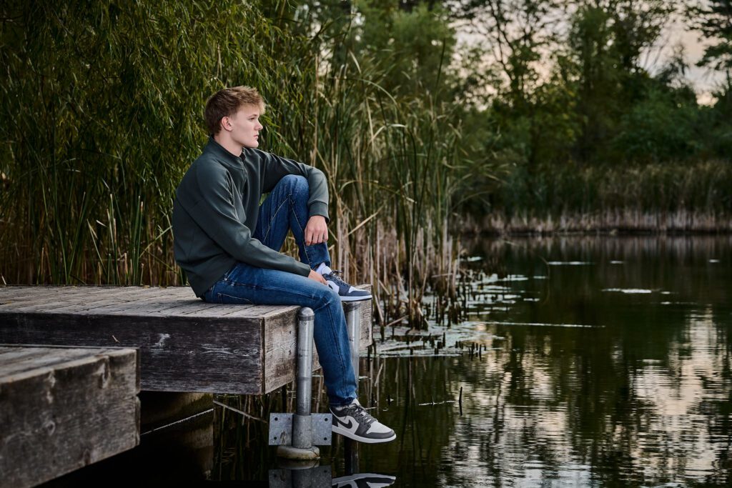 Senior guy sitting on the edge of a dock, looking over the pod, with the pond and grass in the background